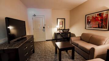 A hotel room with a flat-screen TV on a dark wooden dresser, a beige sofa, a small wooden table, and a framed abstract painting on the wall.