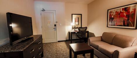 A hotel room with a flat-screen TV on a dark wooden dresser, a beige sofa, a small wooden table, and a framed abstract painting on the wall.