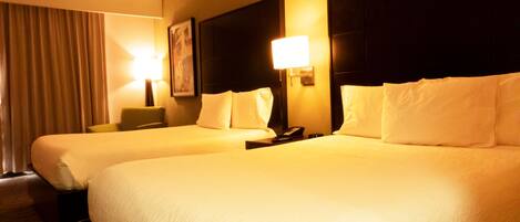 Habitación de hotel con dos camas, cada una con ropa de cama blanca, cabeceros, lámparas de noche y un cuadro en la pared.