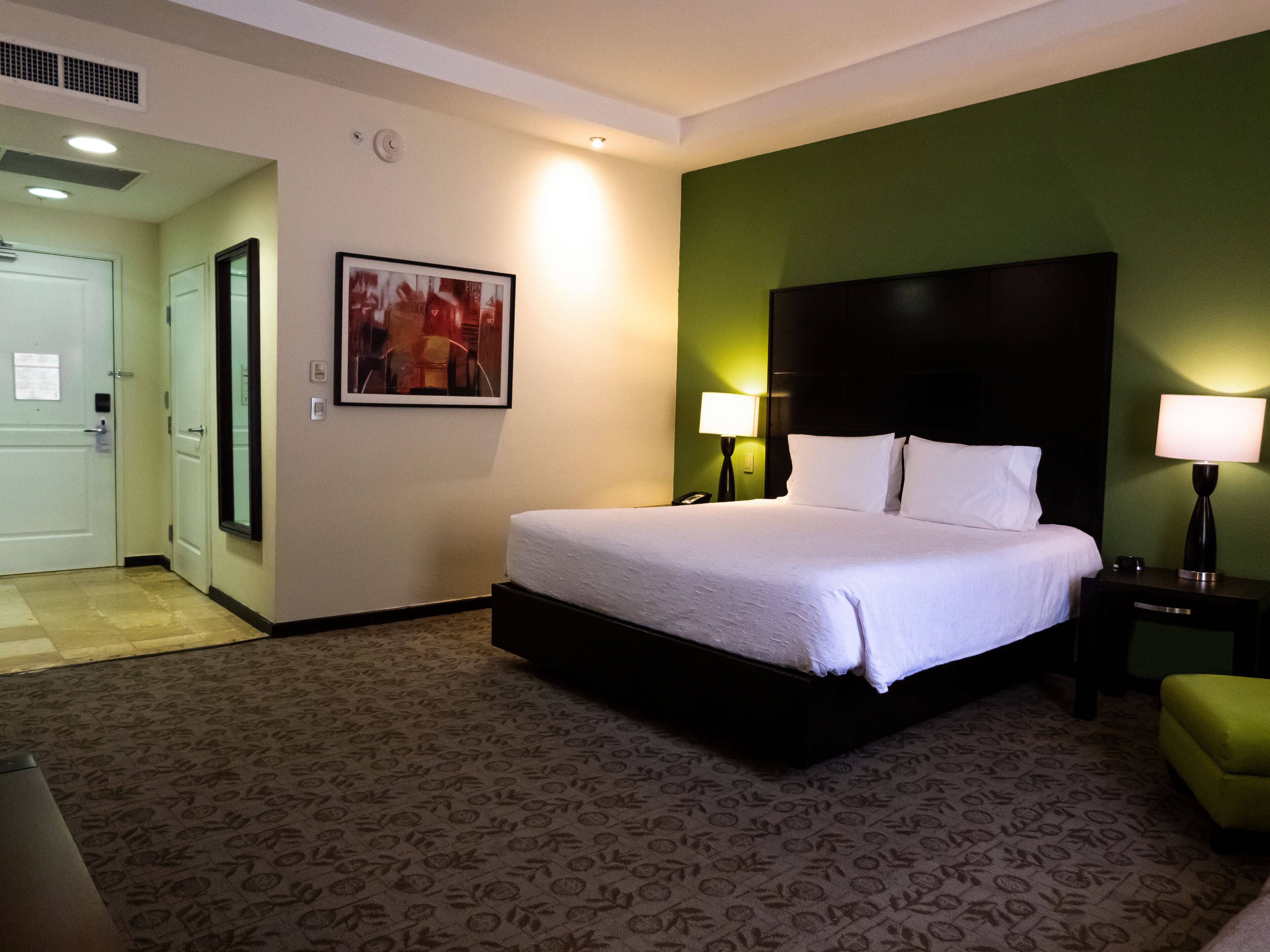 A hotel room with a bed, two bedside lamps, a green accent wall, a framed picture, and a door to the left.