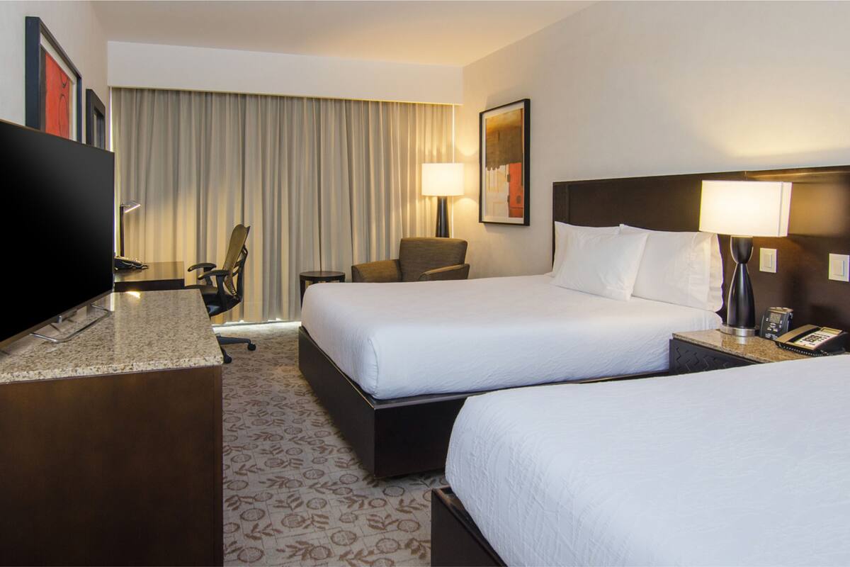 A hotel room with two beds, a desk, a chair, a TV, and a lamp.