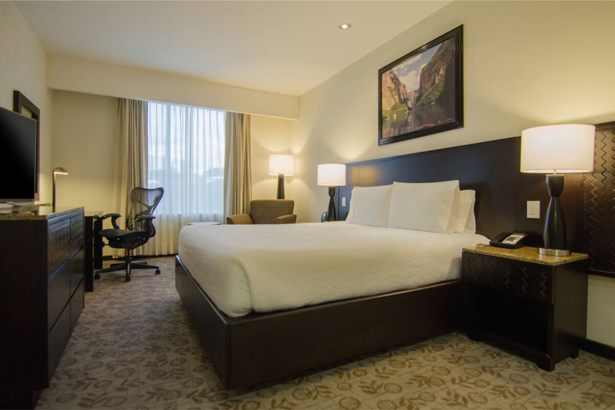 A hotel room with a large bed, a desk with a chair, two bedside lamps, a television, and a painting on the wall.