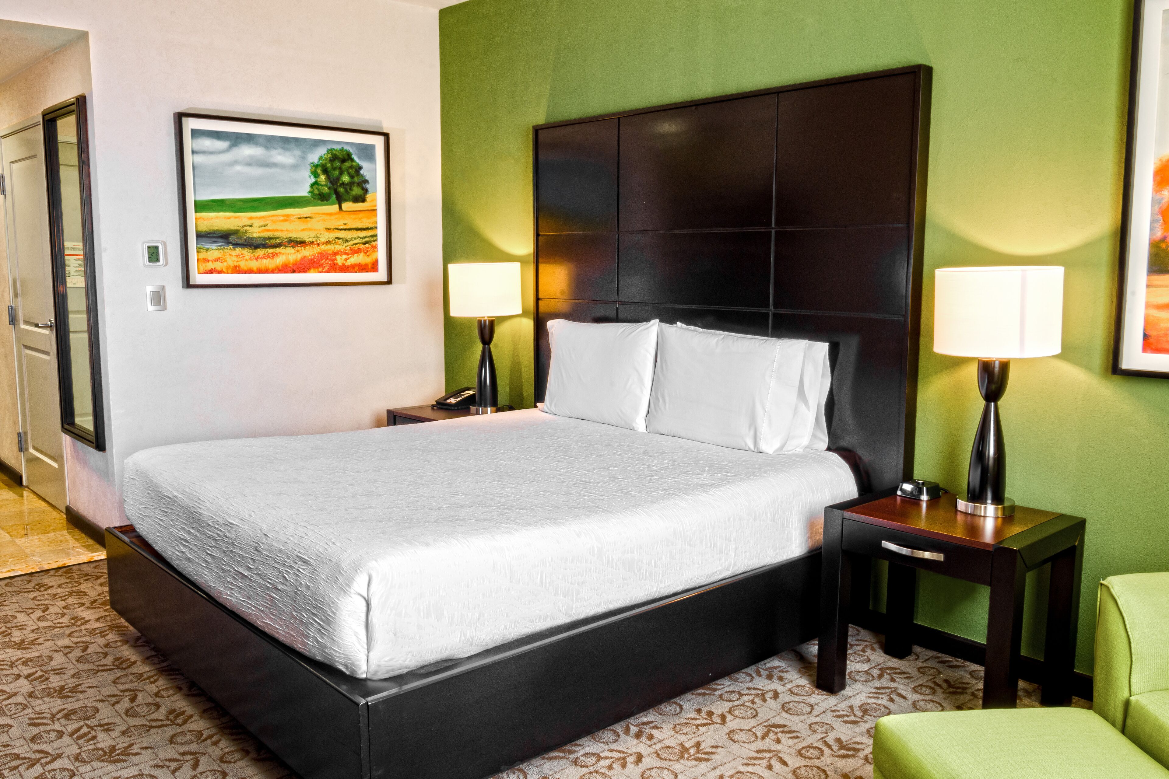 Habitación de hotel con una cama grande, dos mesitas de noche con lámparas, una pared verde decorativa y un cuadro con un paisaje.