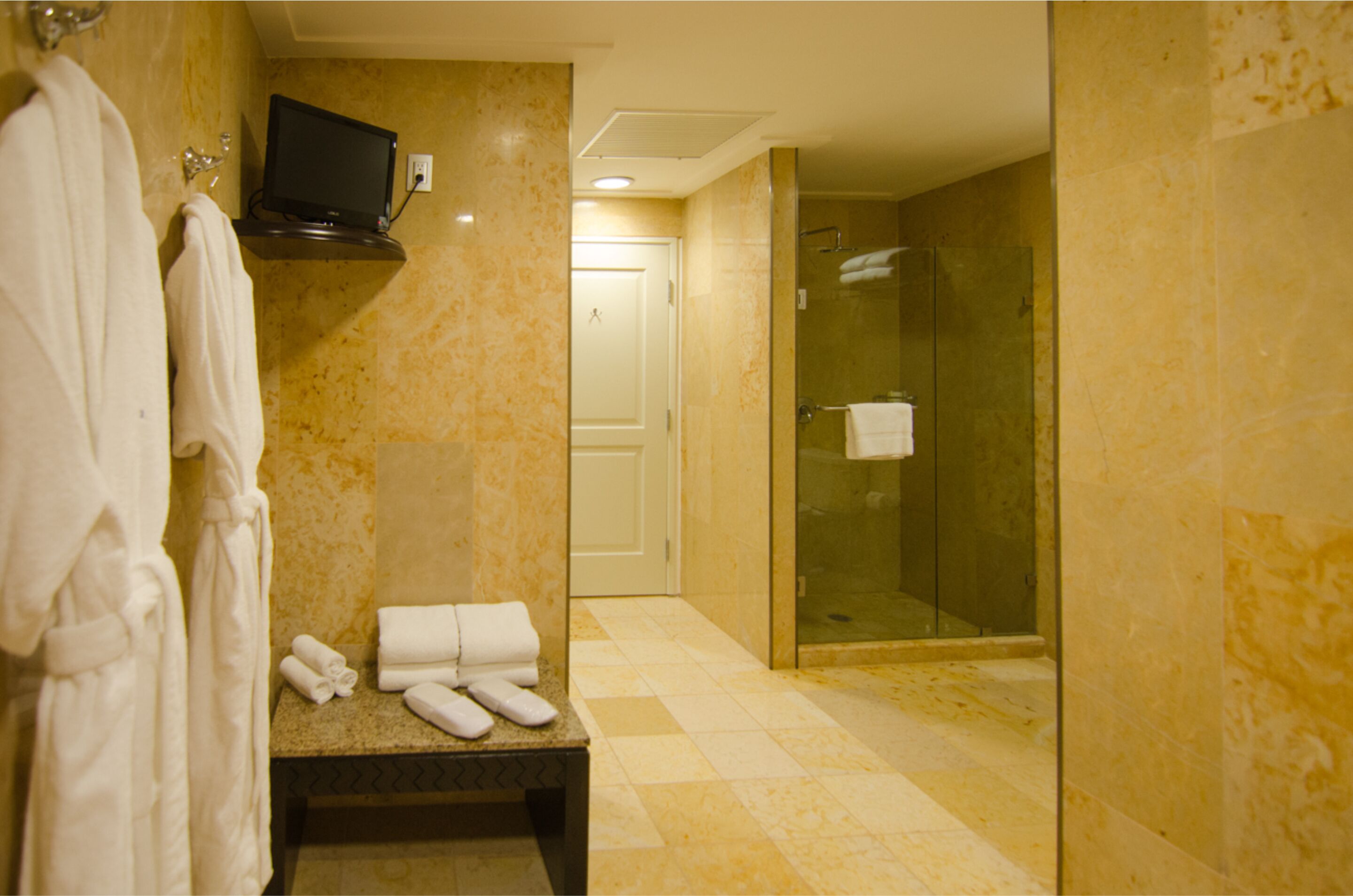 A bathroom with a marble floor, a bench with towels, a glass shower enclosure, and a television mounted on the wall.