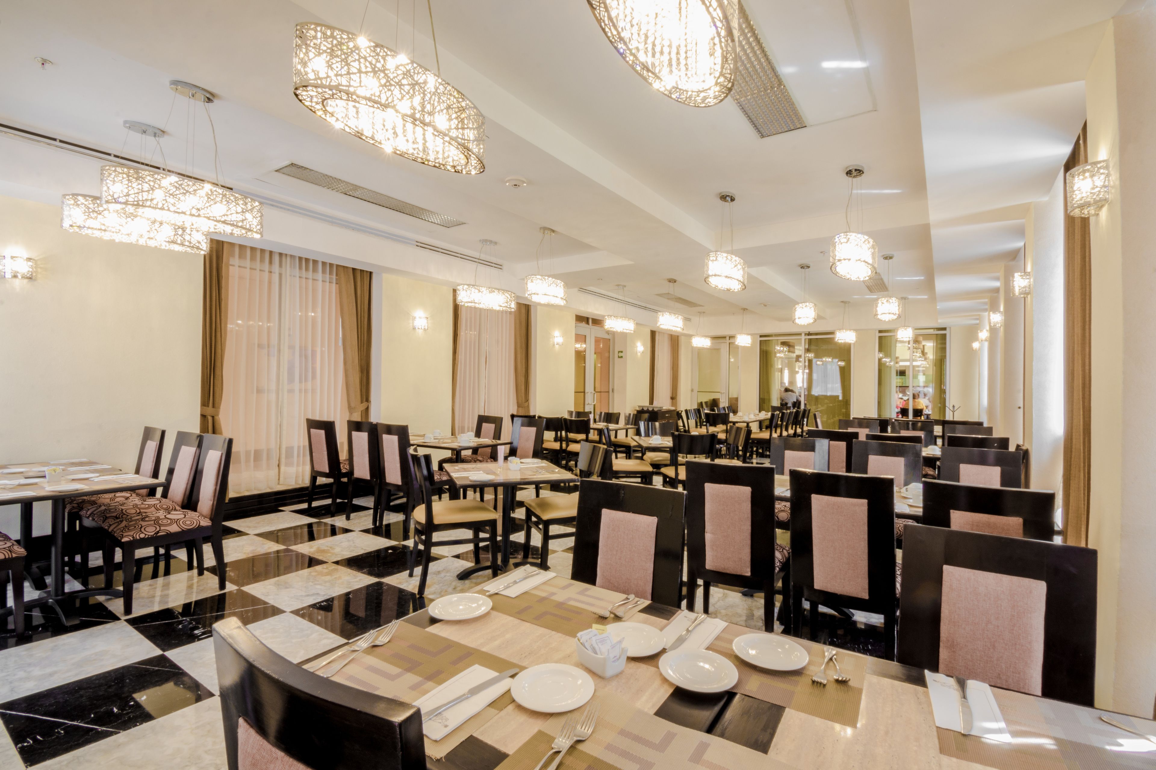A hotel dining area with multiple tables and chairs, chandeliers, and a checkered floor pattern.