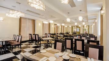 Een restaurantruimte in een hotel met meerdere tafels en stoelen, kroonluchters en een geruit vloertapijt.