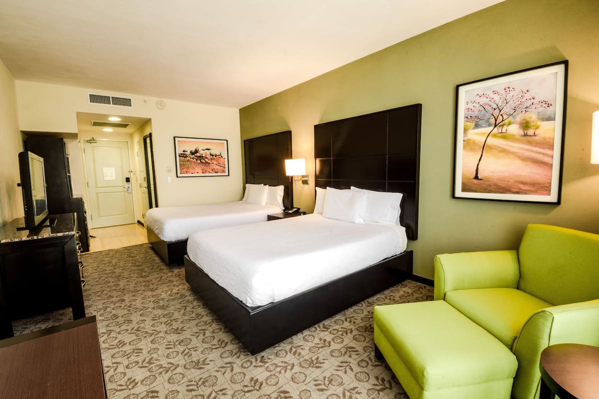 A hotel room with two beds, a desk, a TV, a green armchair, and a painting on the wall.