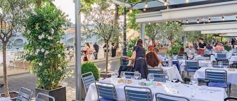 An outdoor dining area with tables and chairs, surrounded by greenery and a view of a bridge.