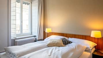 A hotel room with a bed, two bedside lamps, a window with curtains, and a radiator.