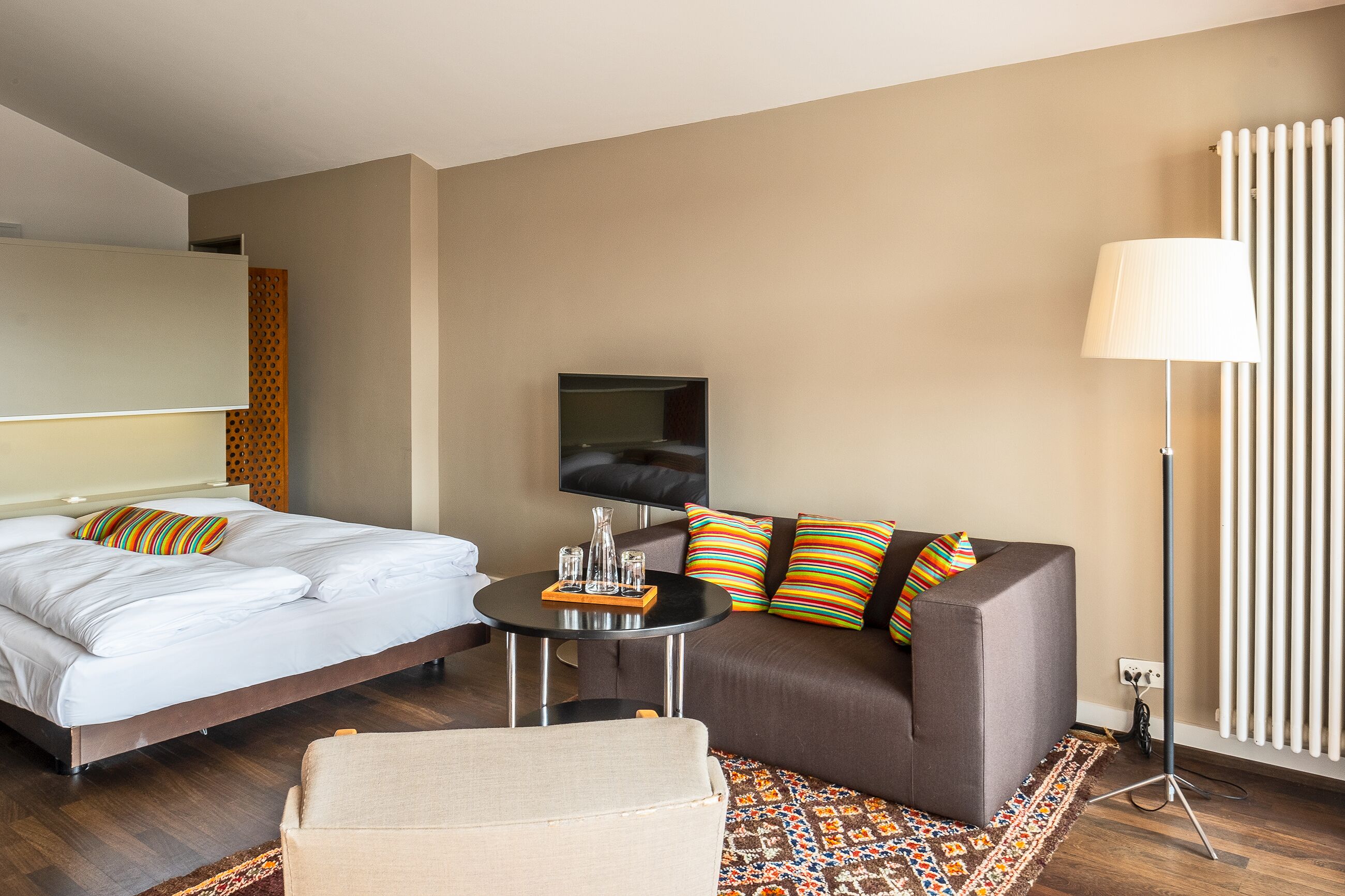 Ein Hotelzimmer mit einem Bett, einem Sofa, einem kleinen Tisch mit Gläsern, einem Fernseher und einer Stehlampe.