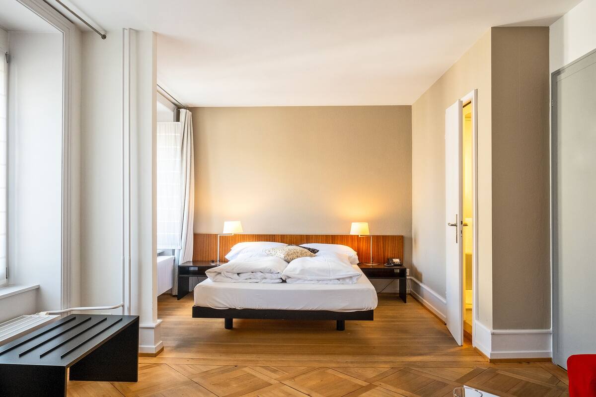 A hotel room with a bed, bedside lamps, a bench, and a view of the hallway.