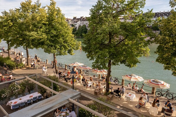 Un café situé au bord de la rivière, avec une terrasse, des vélos à disposition et une vue sur la rivière et les bâtiments en arrière-plan.