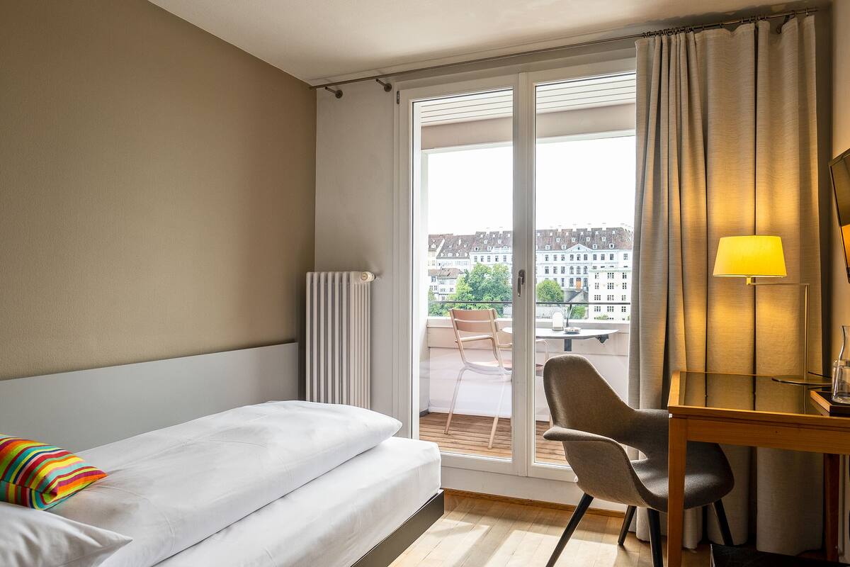 A hotel room with a bed, a desk, a chair, a lamp, and a view of buildings through a window.