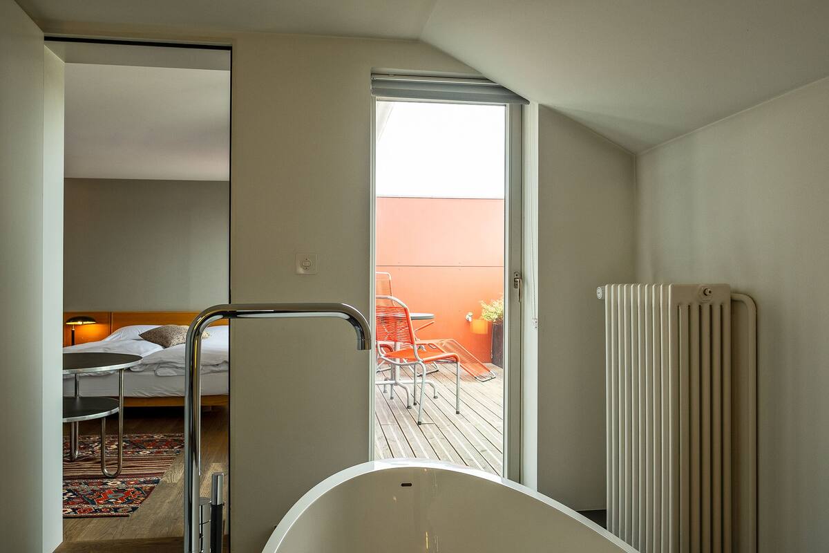 A modern hotel room with a bed, a small table, a chair, a radiator, and a view of an outdoor deck with chairs.