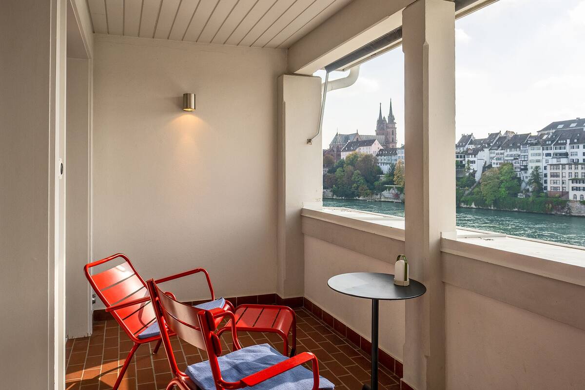 A balcony with red chairs, a small table, and a view of a river and buildings.