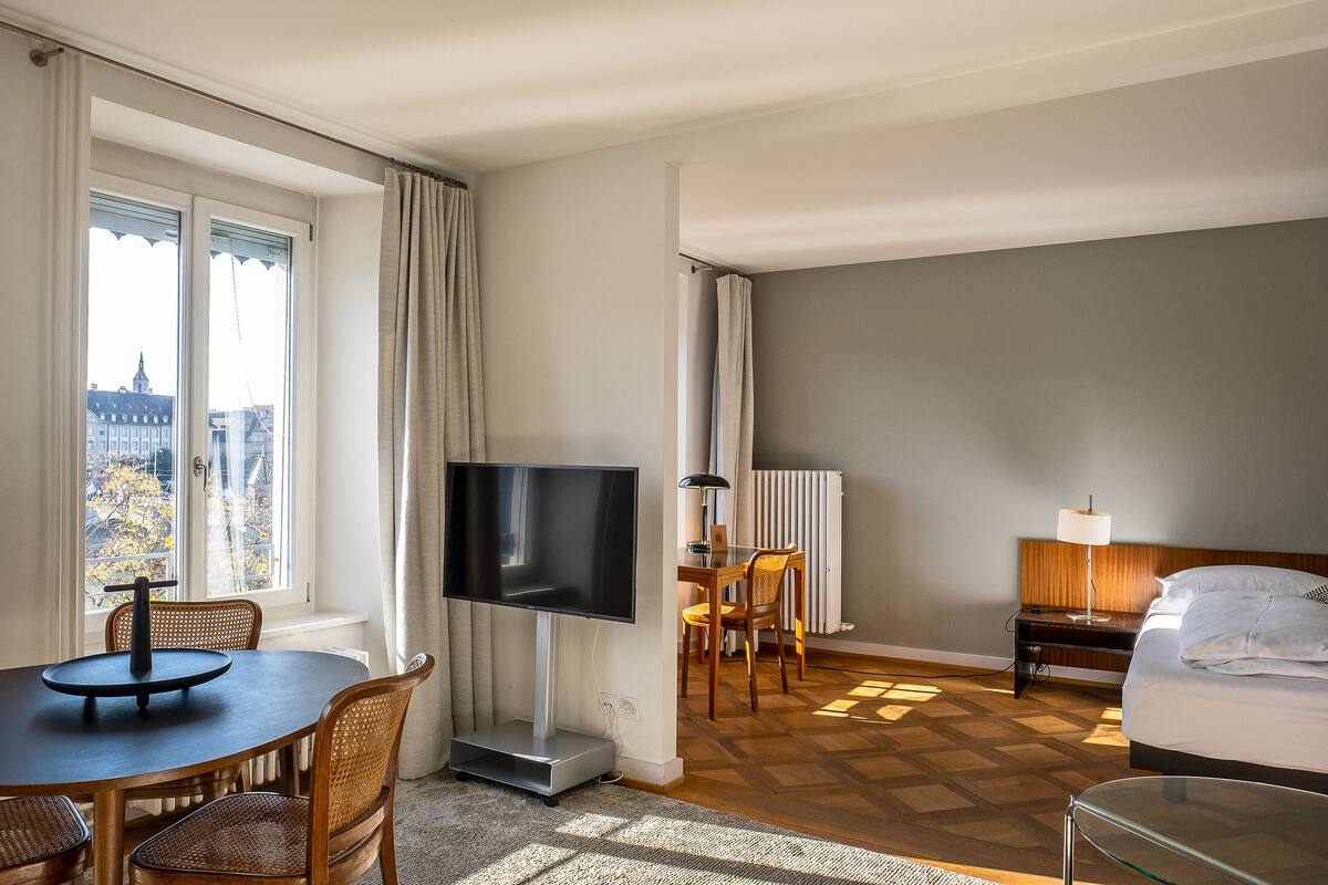 A hotel room with a bed, a desk with a lamp, a TV, and a dining area with chairs.