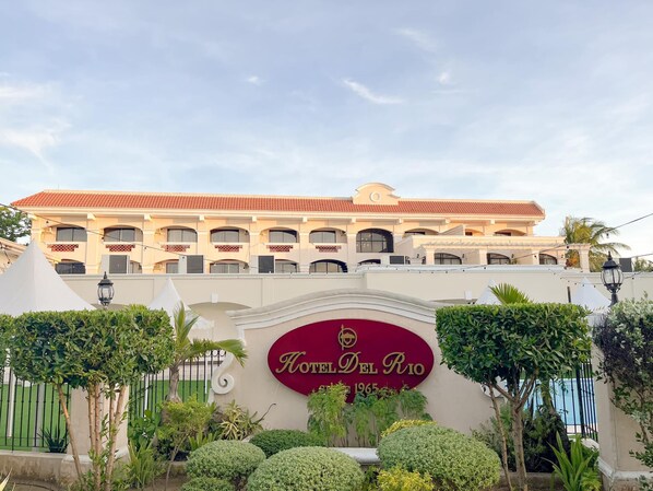 A hotel with a red and white facade, a sign reading "Hotel Del Rio," and a pool area in the background.