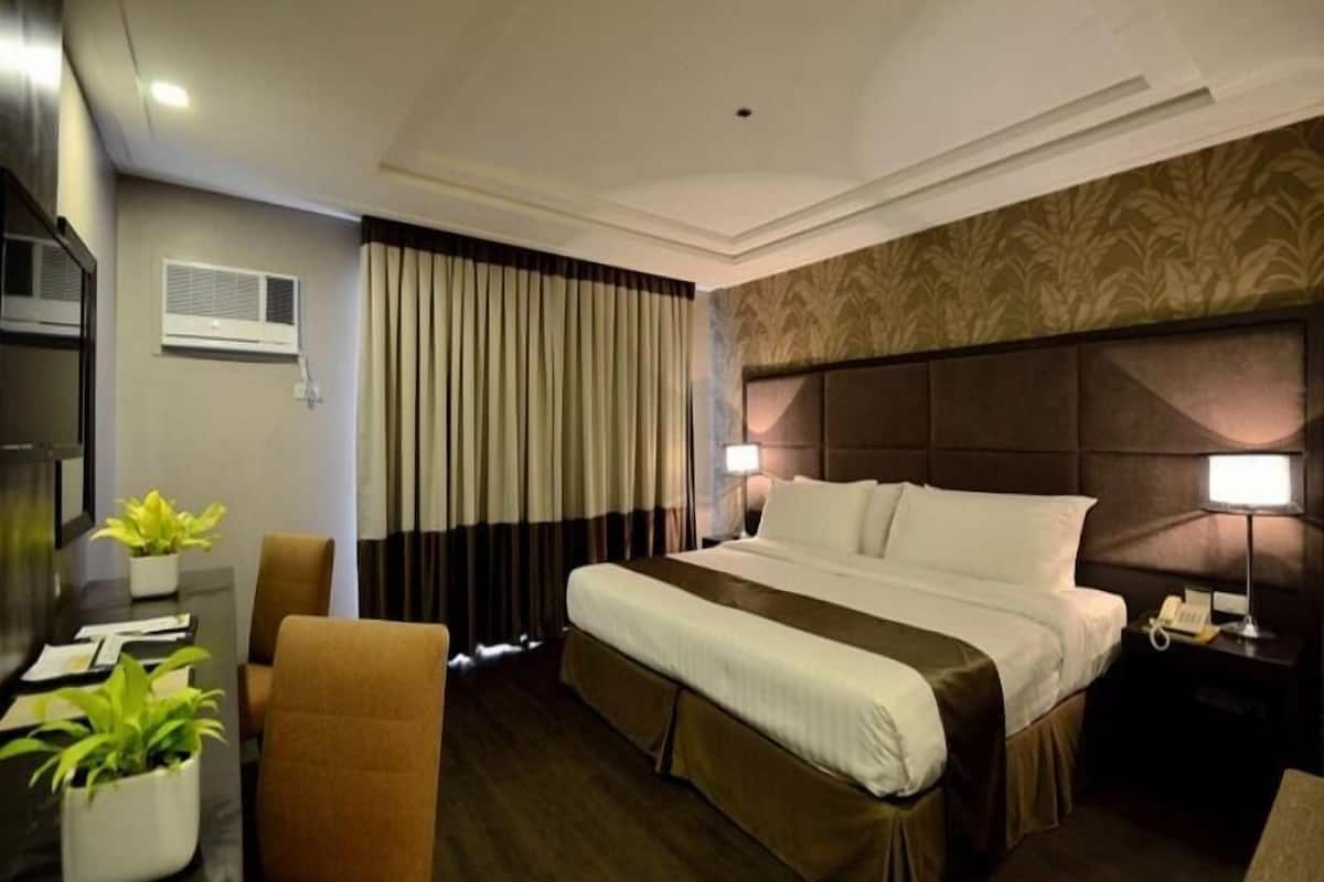 A hotel room with a bed, a desk with a chair, a potted plant, a nightstand with a lamp, and a window with curtains.