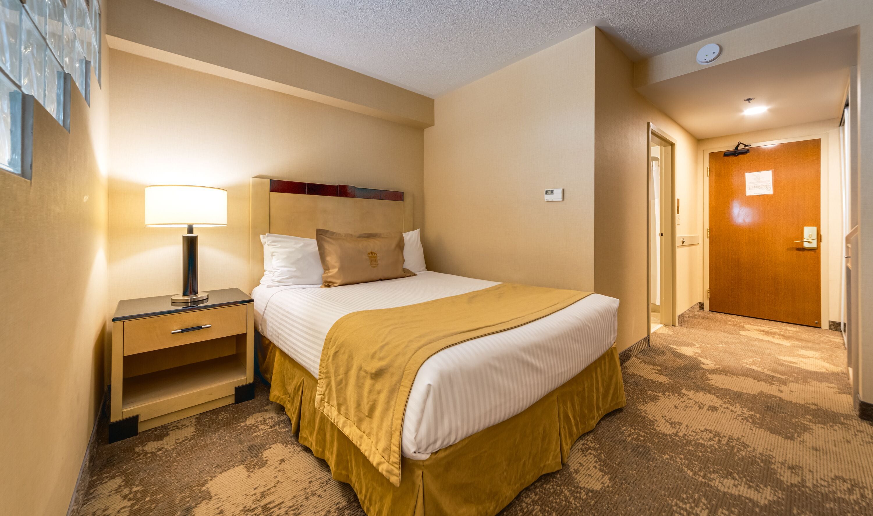 Una habitación de hotel con cama, mesita de noche, lámpara y una puerta que da a otra estancia.