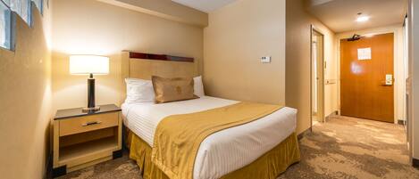 A hotel room with a bed, bedside table, lamp, and a door leading to another room.