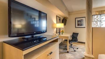 A hotel room with a flat-screen TV on a wooden dresser, a desk with a chair, a lamp, and framed pictures on the wall.