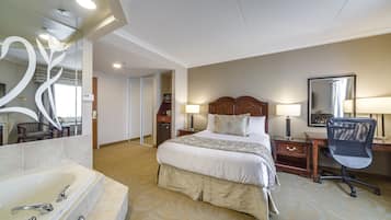 Quarto de hotel com uma cama grande, uma escrivaninha, uma cadeira, luminária de mesa, mesa de cabeceira, espelho e banheiro com banheira e pia.