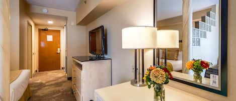 Una habitación de hotel con una cama, un escritorio con lámpara, un espejo y un florero con flores.