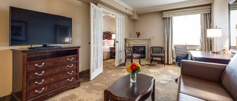 A hotel room with a flat-screen TV, a wooden dresser, a sofa, a coffee table, a fireplace, and a bed with a nightstand.