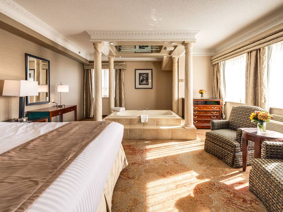 A hotel room with a large bed, a sitting area with a chair, a small table, a desk, a mirror, a lamp, a bathtub, and a dresser.