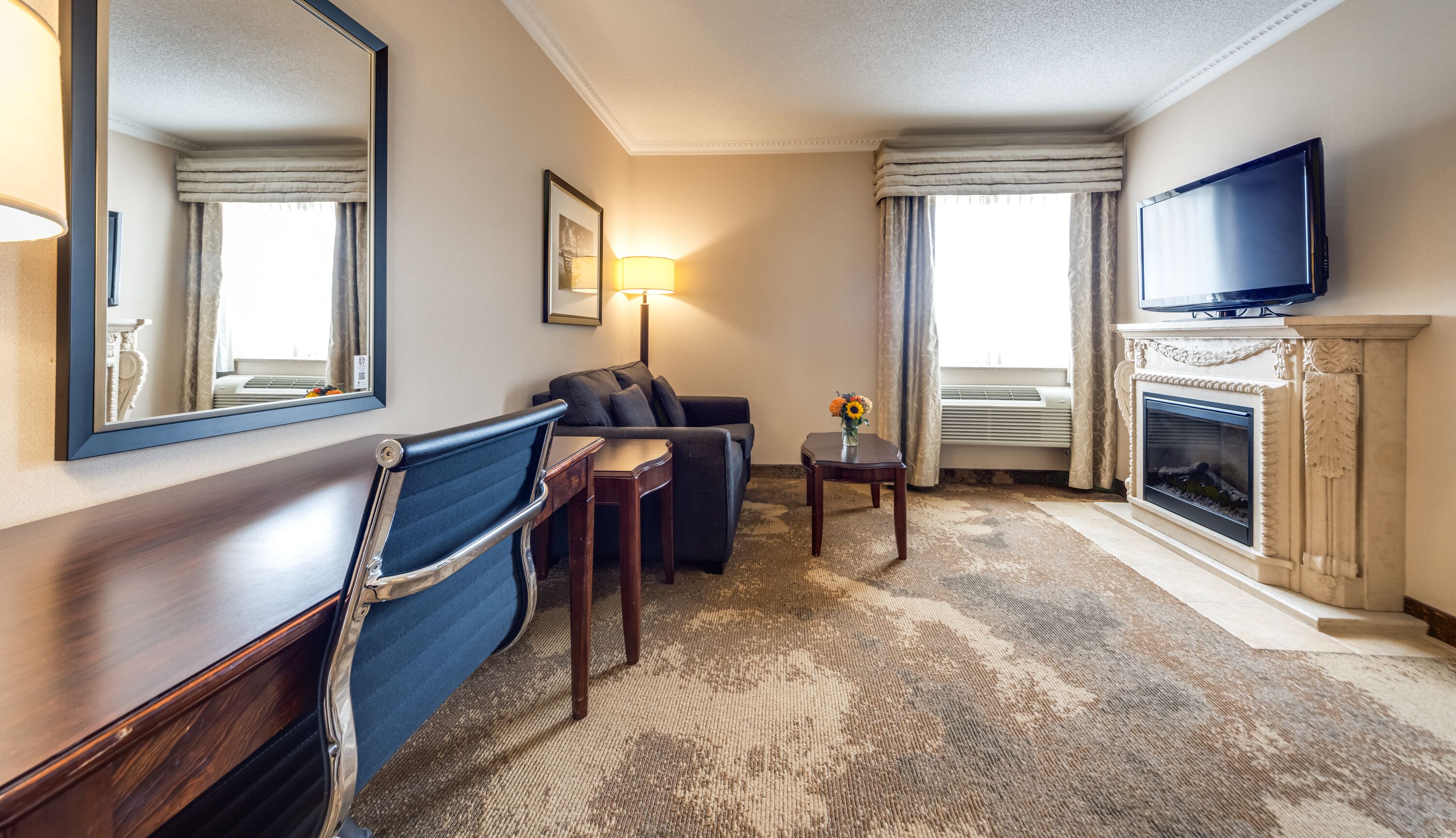 A hotel room with a sofa, a small table, a lamp, a TV, and a fireplace.