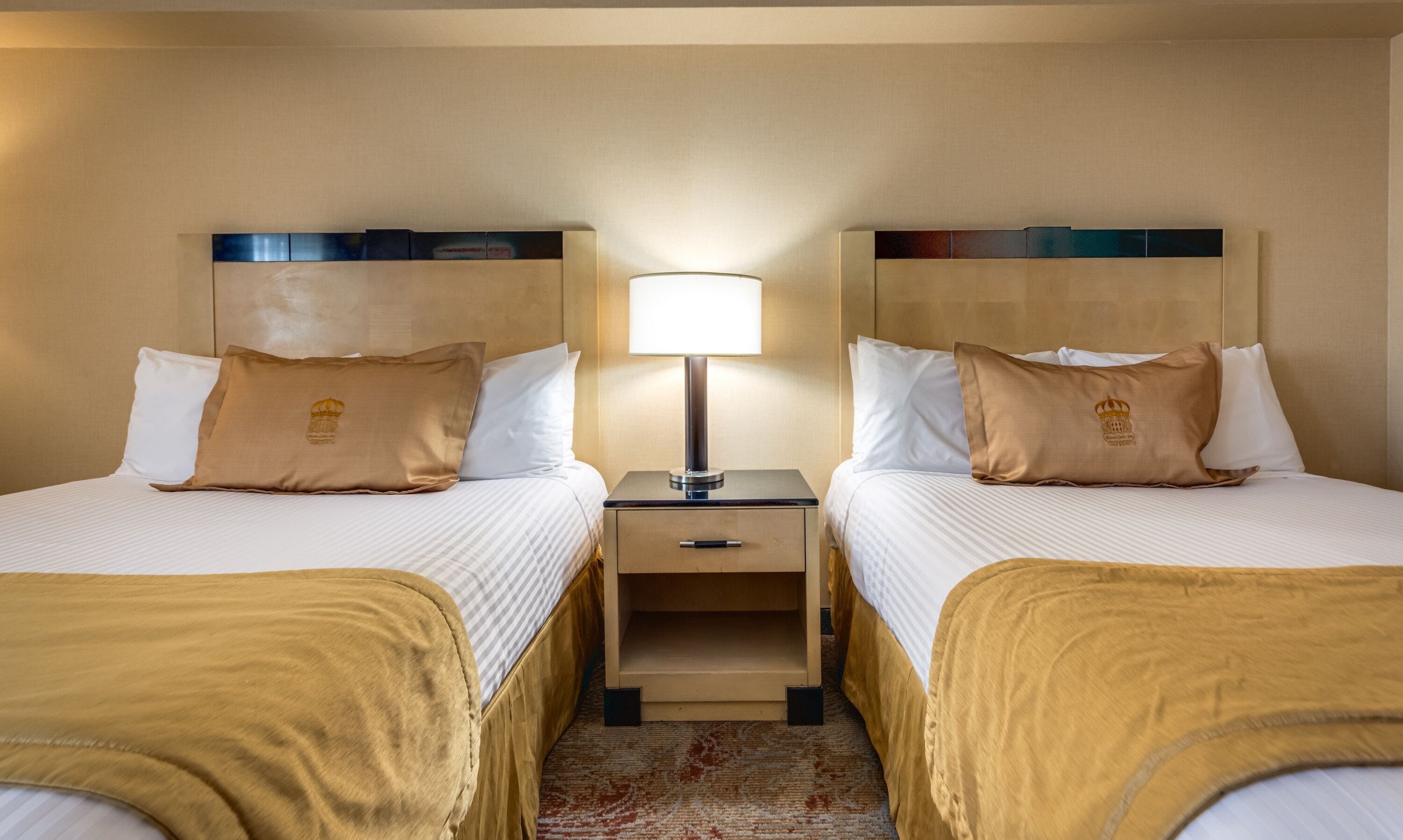 Une chambre d’hôtel avec deux lits, chacun doté d’une tête de lit marron, d’une table de chevet avec une lampe et d’un petit tiroir.