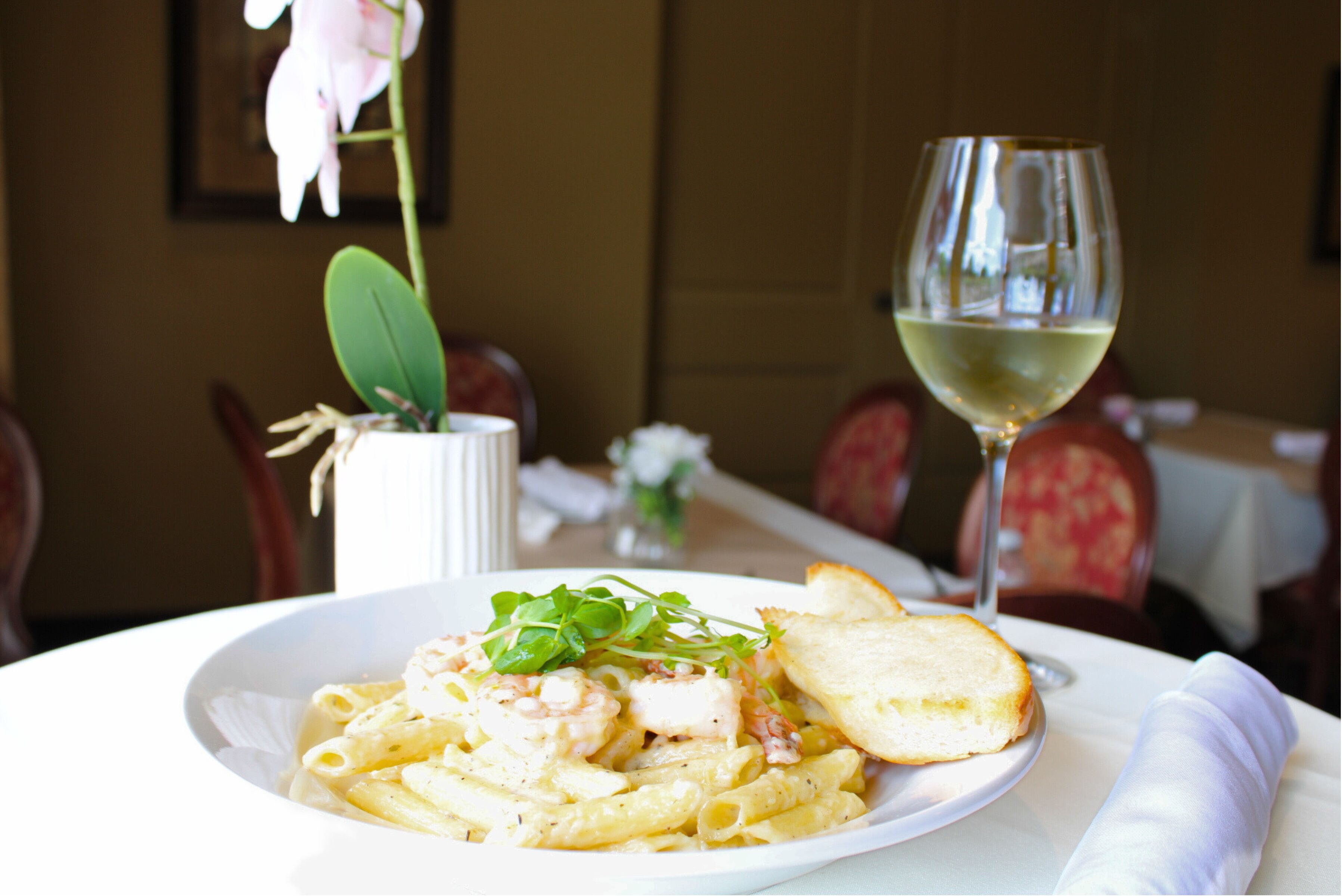 A plate of pasta with shrimp and bread, a glass of white wine, and a vase with flowers on a table.