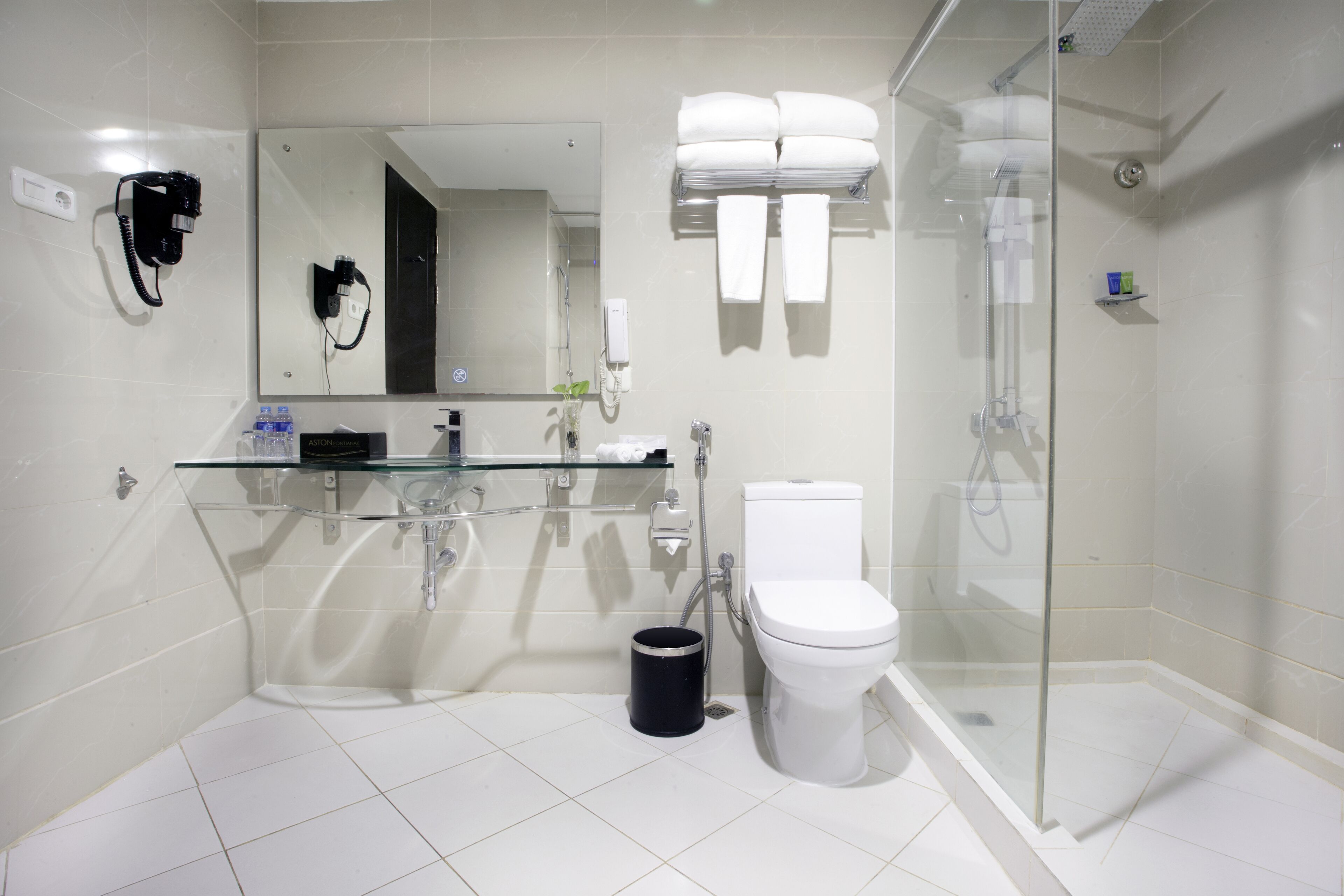 A modern bathroom with a glass shower enclosure, a white toilet, a glass sink, and a hairdryer mounted on the wall.