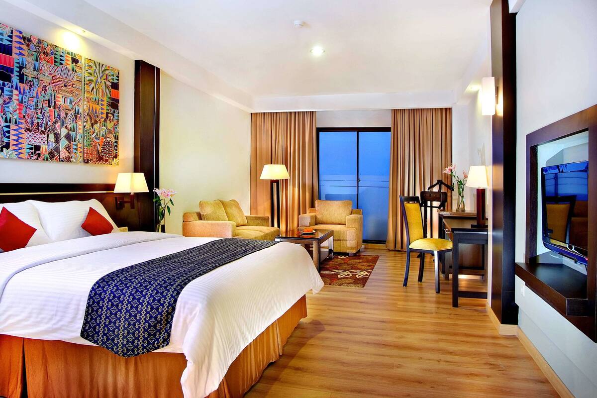 A hotel room with a large bed, two armchairs, a desk, a TV, and a colorful abstract painting on the wall.