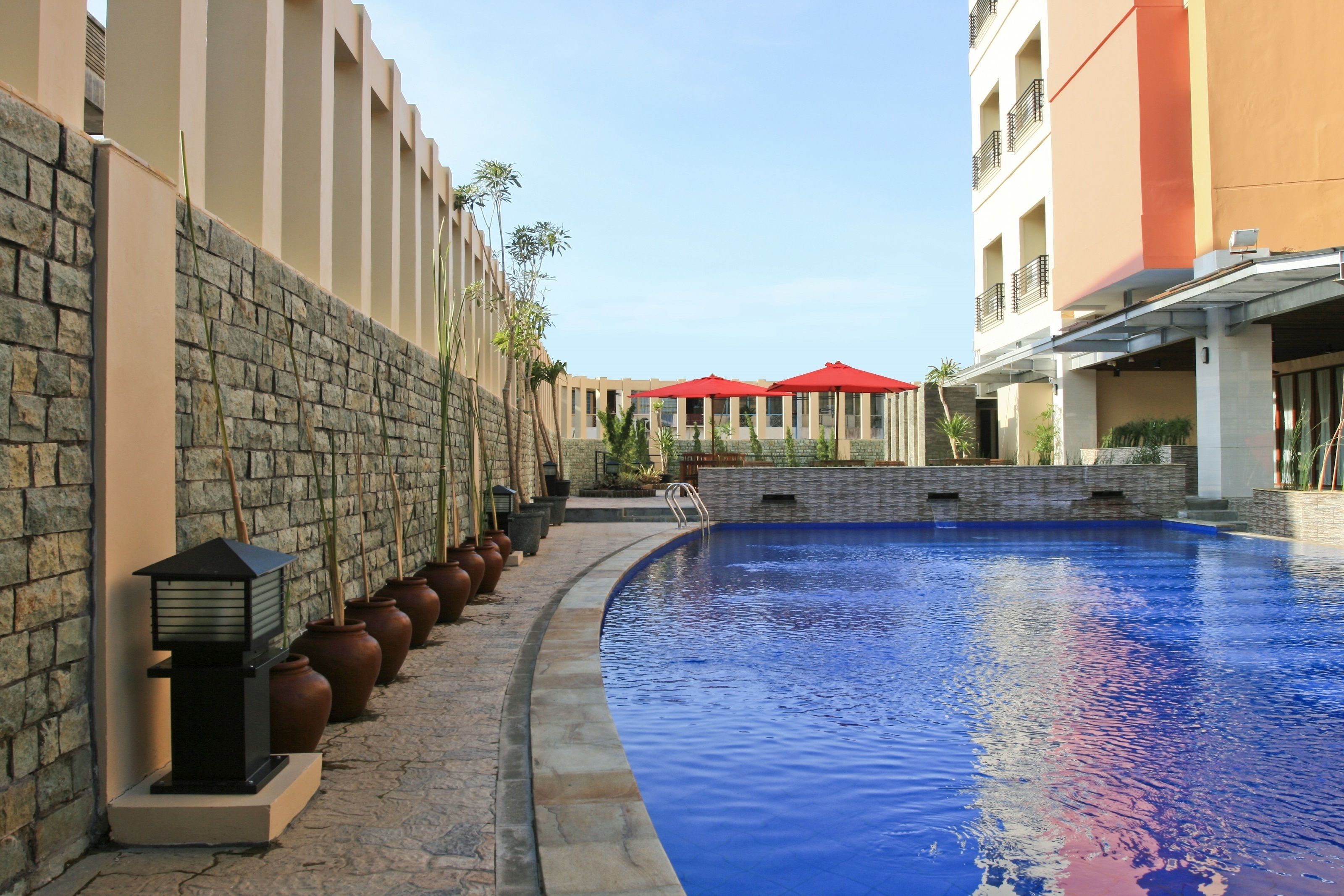 A swimming pool with a stone wall and potted plants on one side, and a building with balconies and red umbrellas on the other.