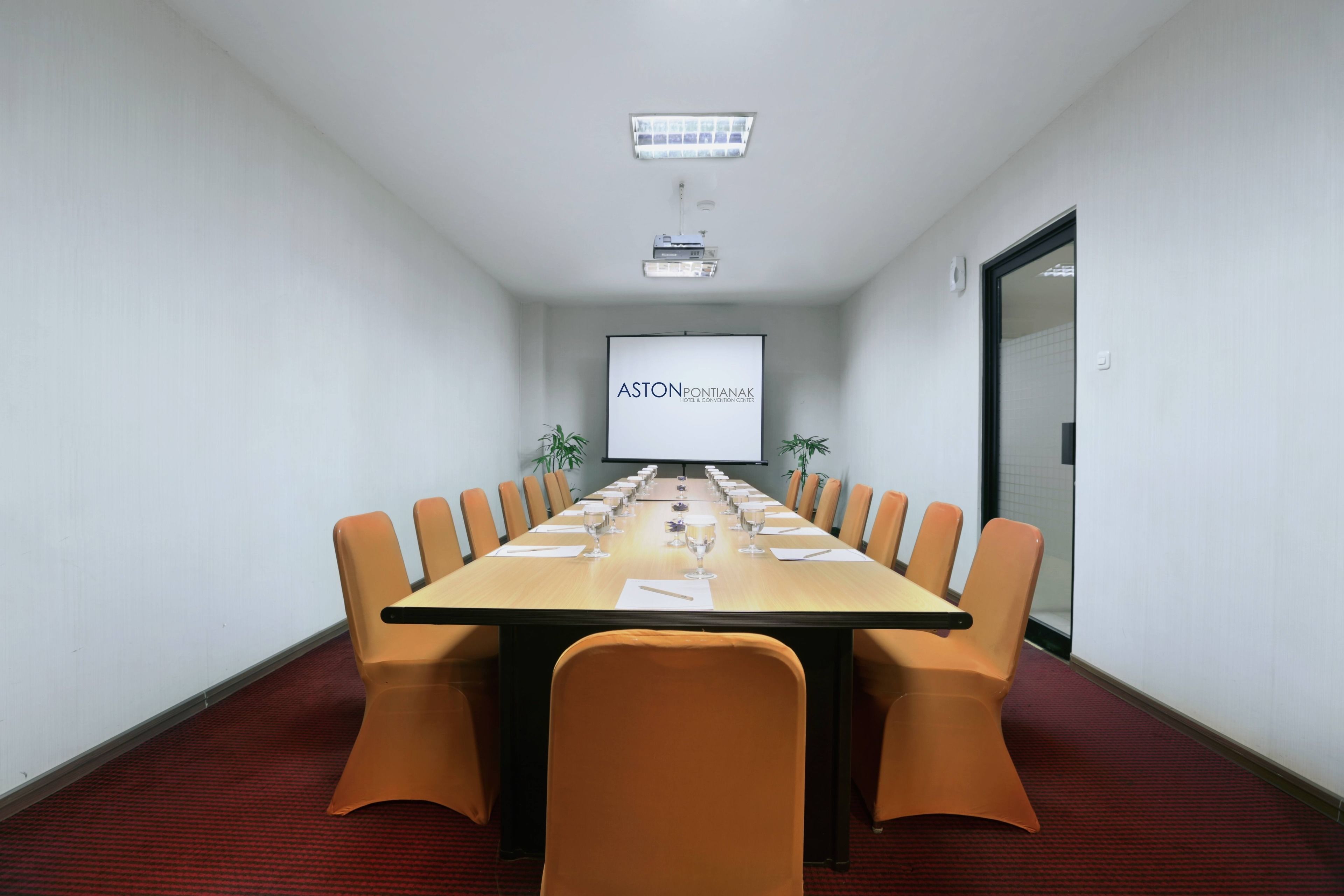 A conference room with a long table, orange chairs, a projector screen, and a red carpeted floor.