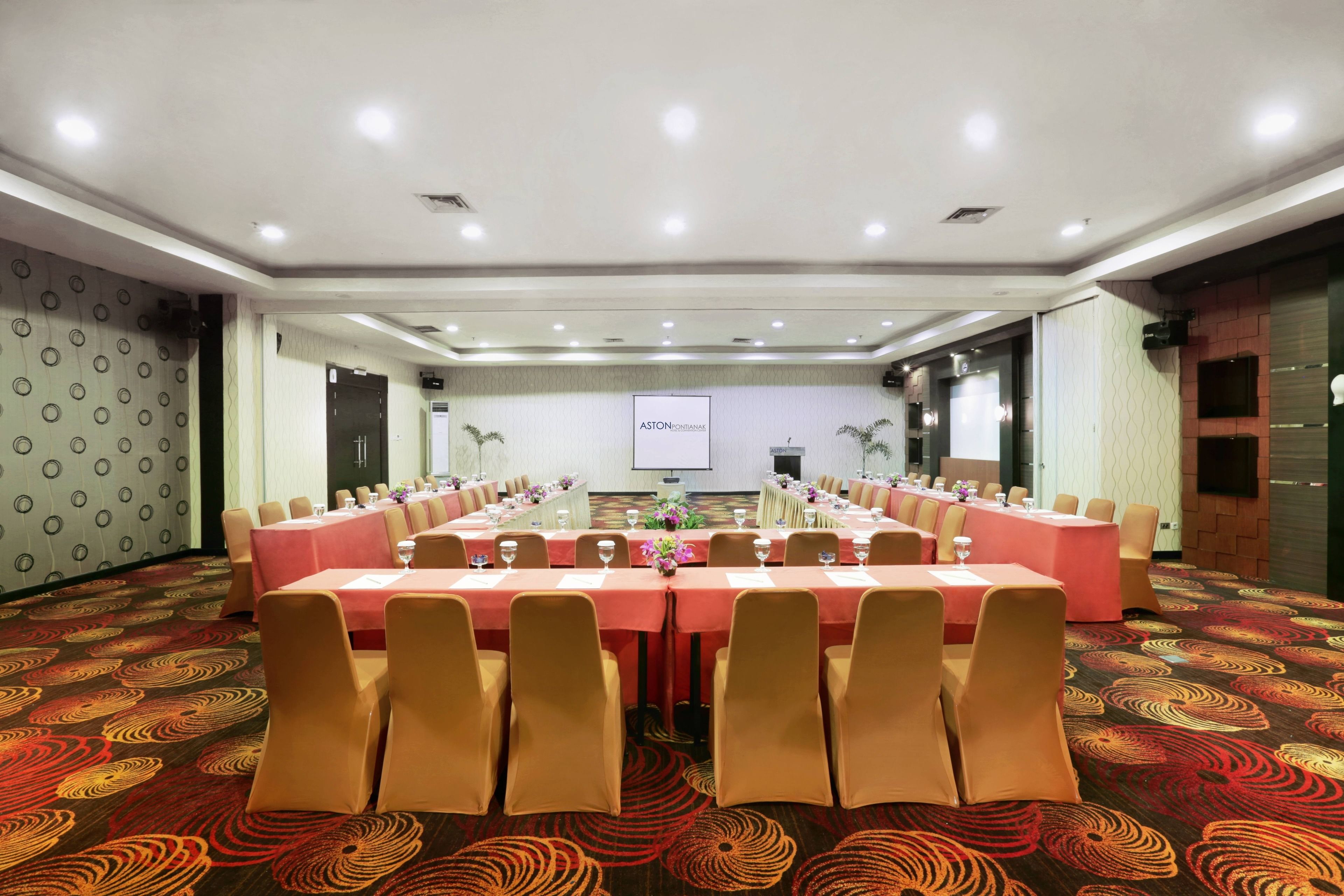 A conference room with a long table set for a meeting, surrounded by chairs, with a projector screen and a floral arrangement on the wall.