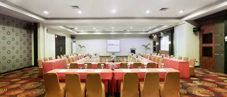 A conference room with a long table set for a meeting, surrounded by chairs, with a projector screen and a floral arrangement on the wall.