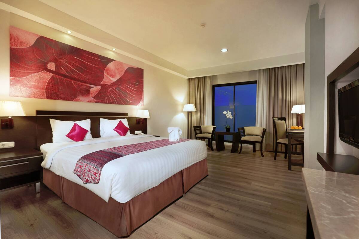 A hotel room with a large bed, two bedside tables, a TV, a desk, and a large window.
