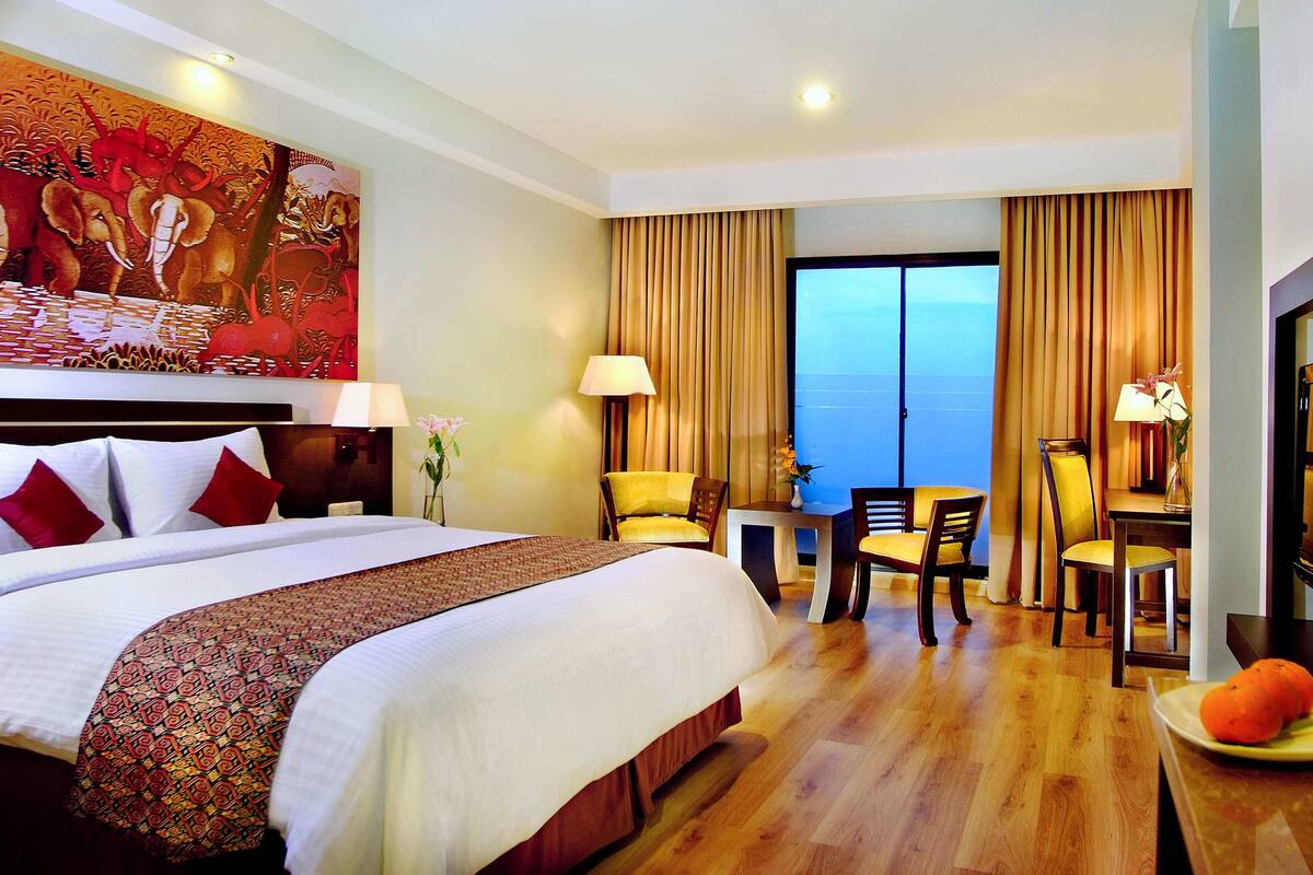 A hotel room with a large bed, a desk, two chairs, a TV, and a view of the sea.