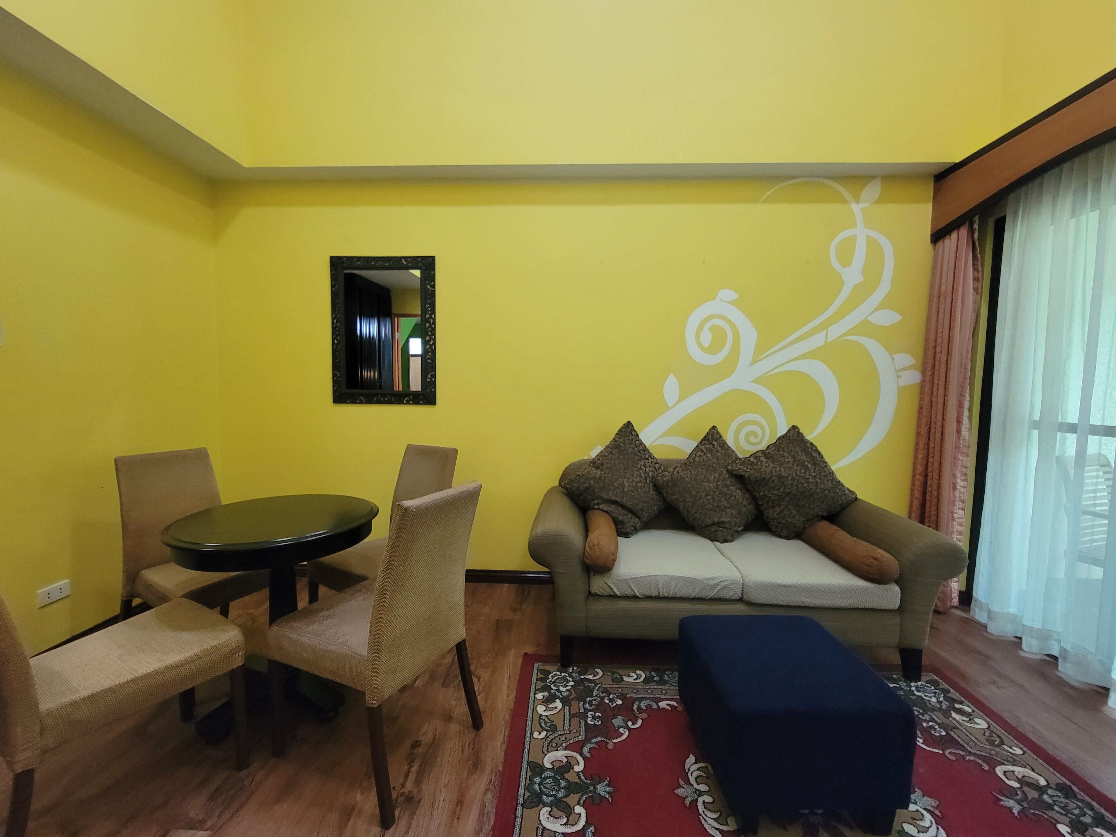 Una sala de estar con una pared amarilla, una mesa redonda, dos sillas, un sofá con cojines, un reposapiés azul y una alfombra estampada.