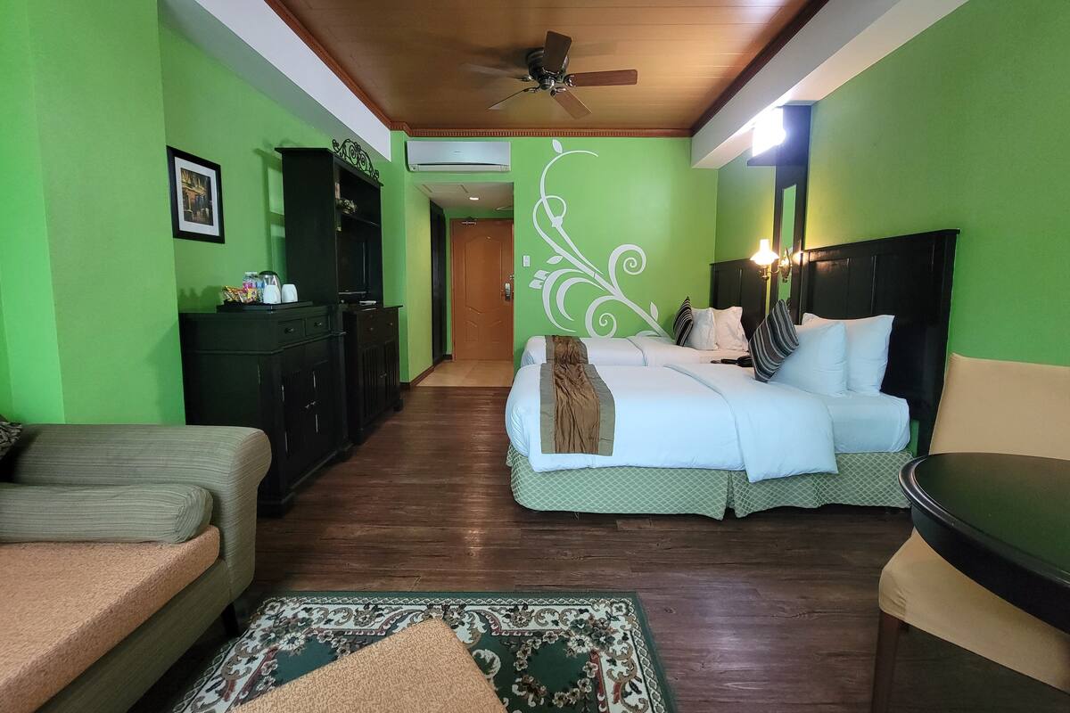 A hotel room with a green wall, a ceiling fan, a bed with white bedding, a wooden floor, and a small seating area.