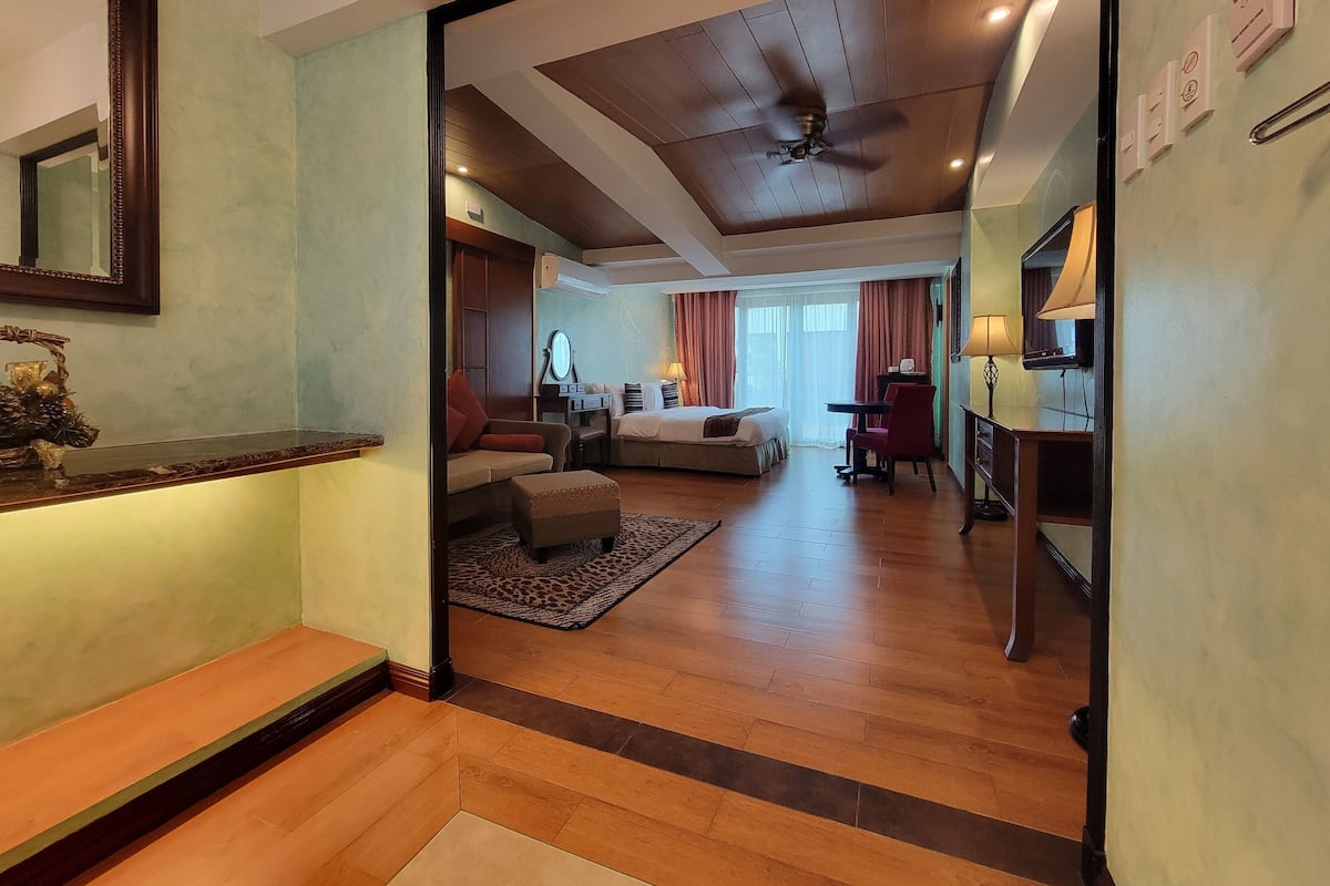 A hotel room with a wooden floor, a bed, a desk, a chair, a TV, and a ceiling fan.