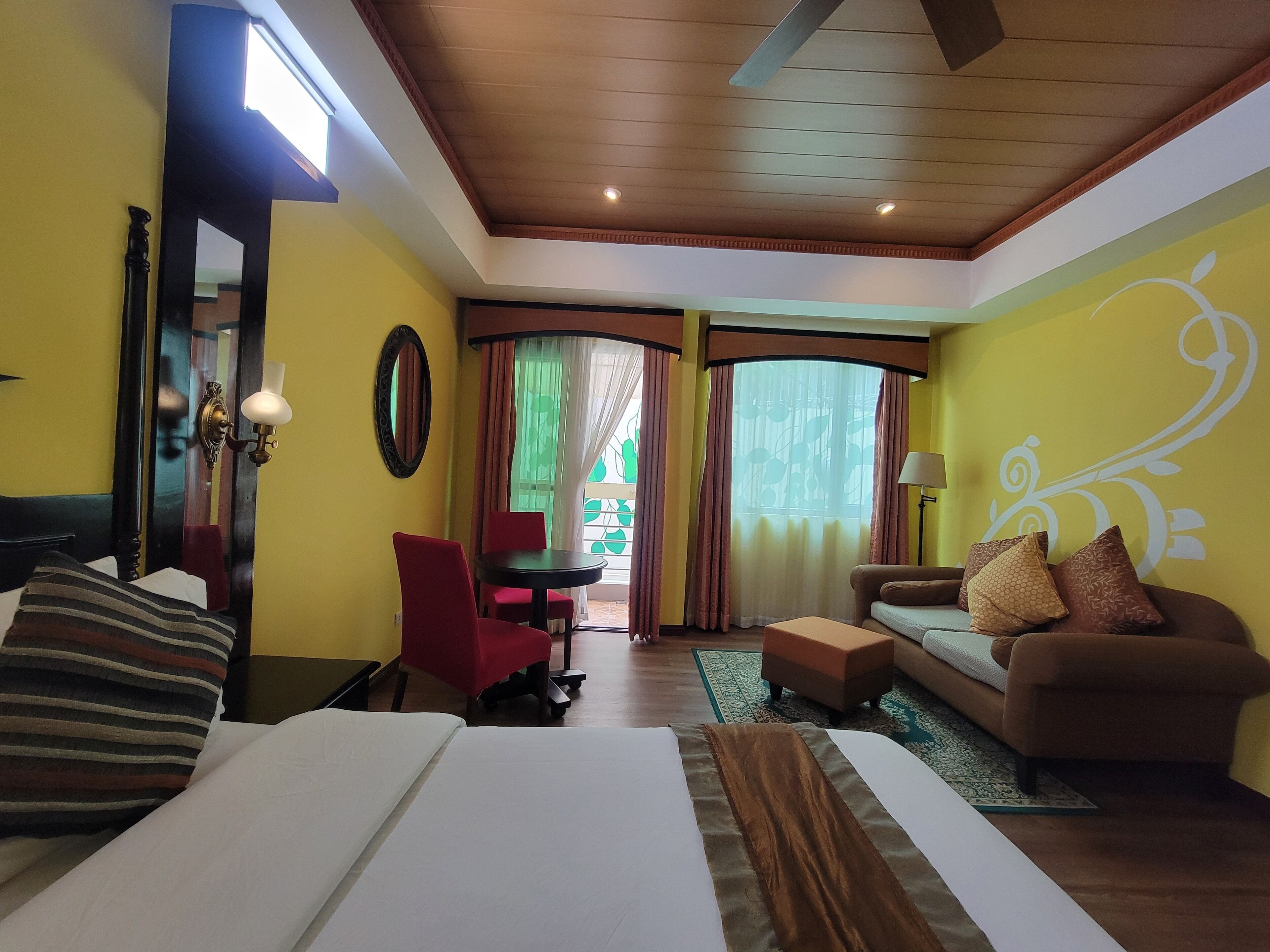 Habitación de hotel con cama, sofá, una mesita y una silla. Cuenta con una ventana con cortinas, un espejo y una lámpara.