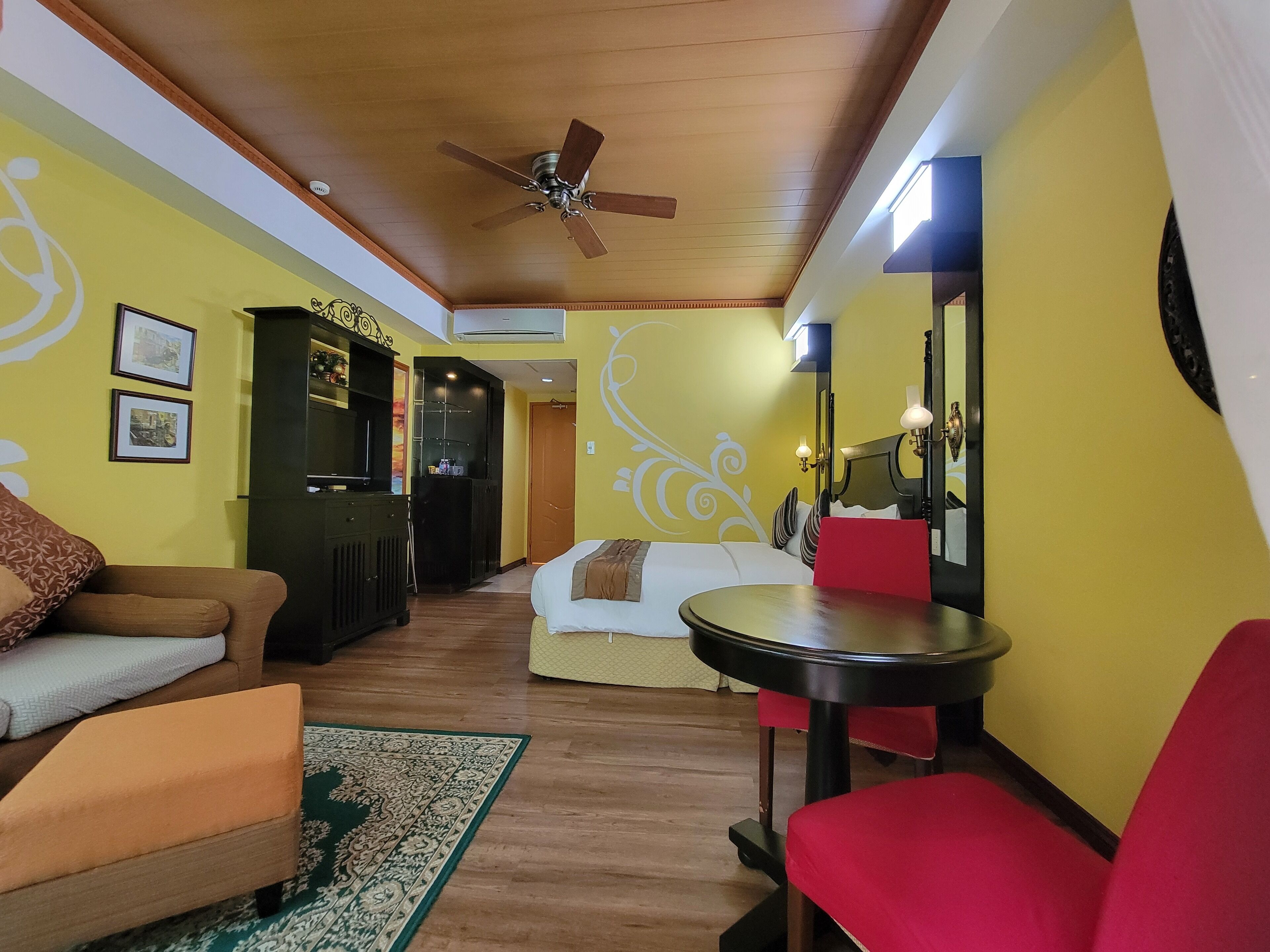 A hotel room with a bed, a small table, a red chair, a sofa, a TV, and a ceiling fan.