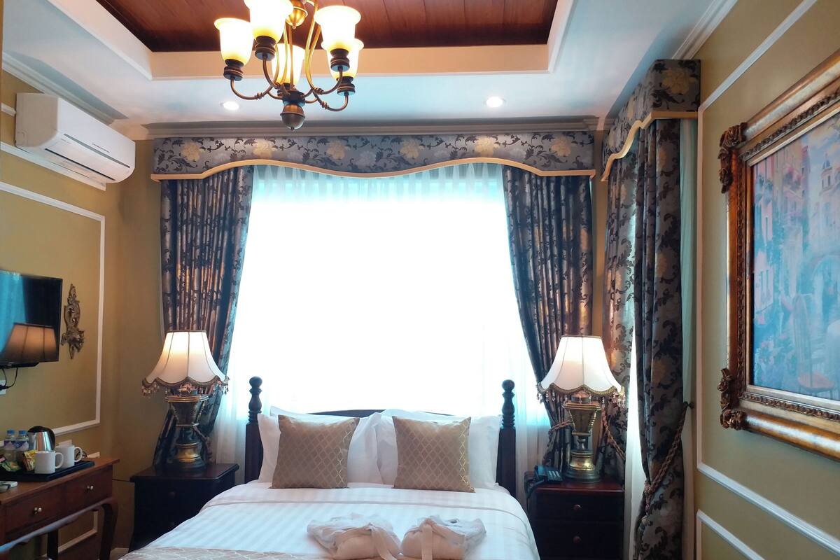 A hotel room with a large bed, two bedside tables, a chandelier, curtains, and a painting on the wall.