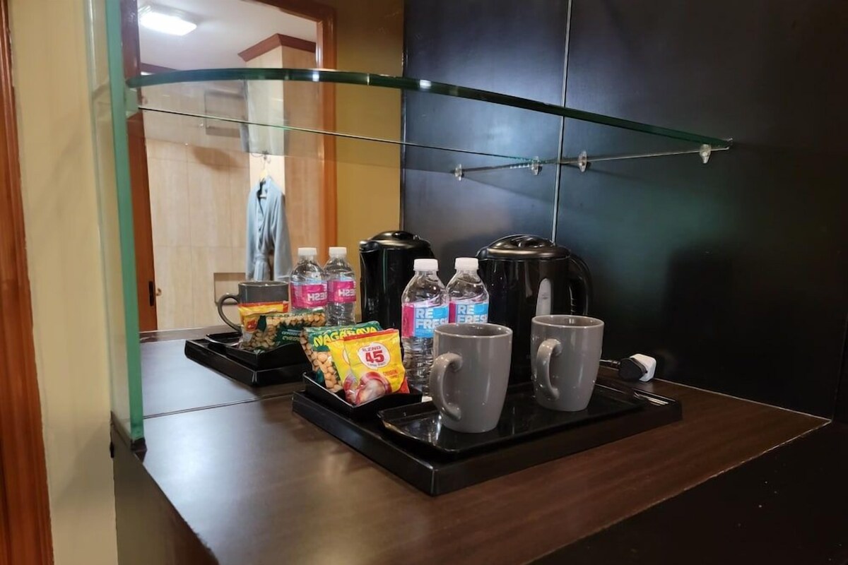 Habitación de hotel con un separador de vidrio, una mesa de madera con una bandeja que contiene aperitivos, agua embotellada y dos tazas.