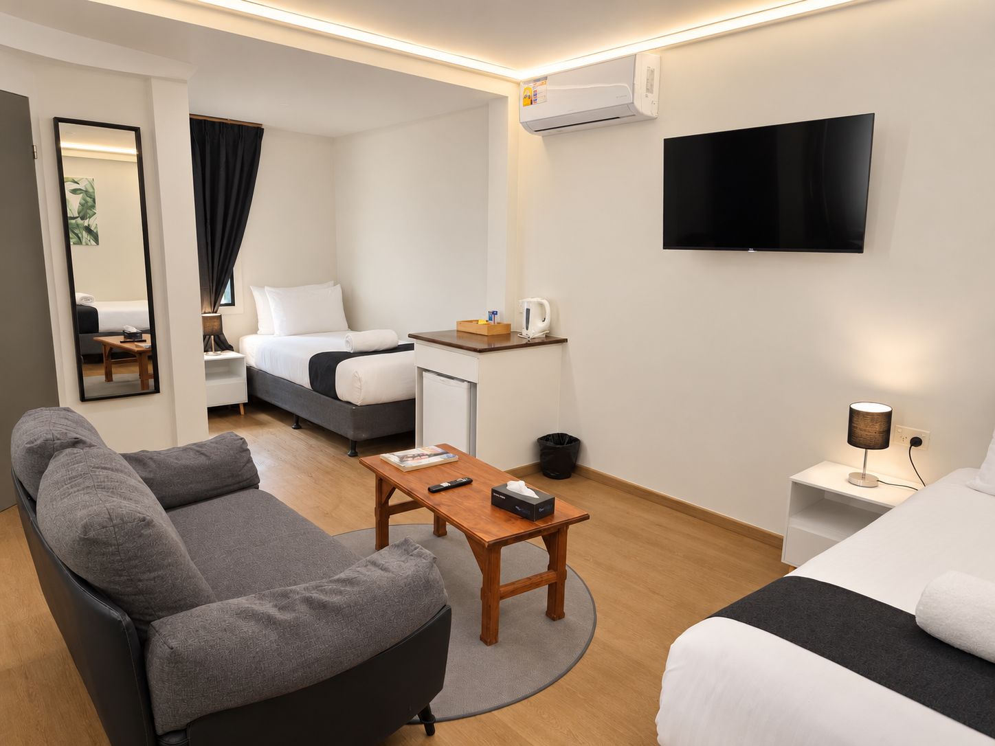 A modern hotel room with a sofa, a bed, a desk, a TV, and a small kitchenette.