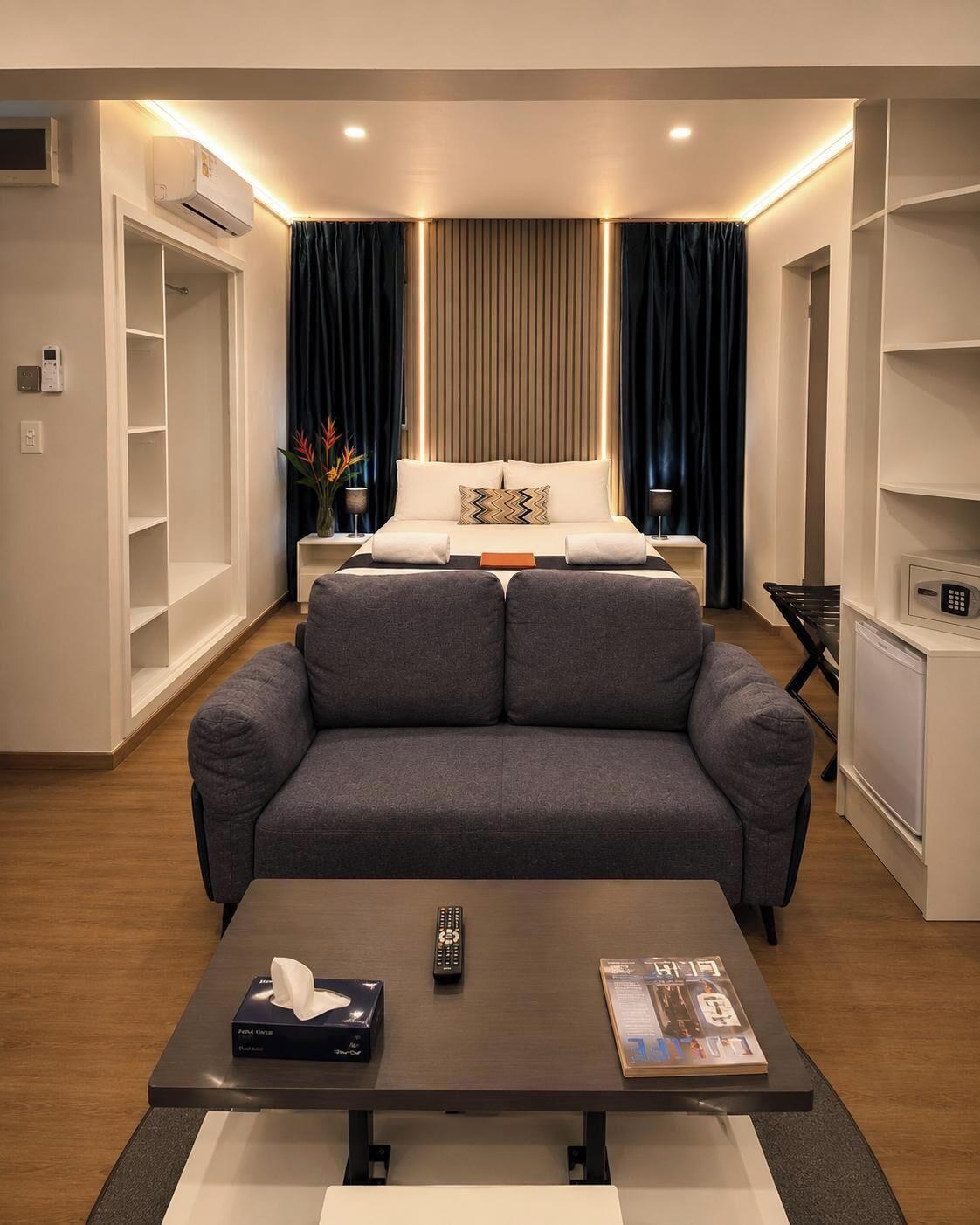 A modern hotel room with a sofa bed, a coffee table with remote controls and a book, a bedside table with a lamp, and a built-in air conditioning unit.