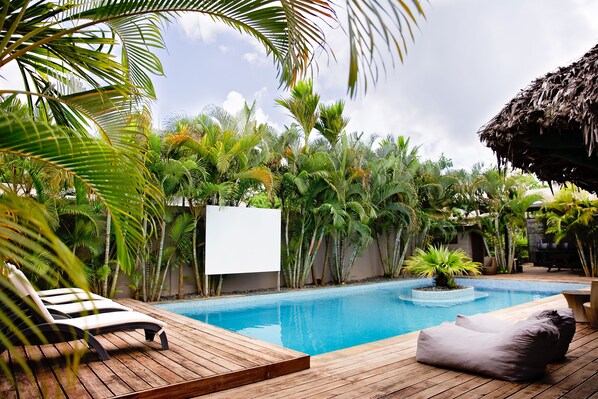 Terraza de madera con sillas de descanso y piscina rodeada de plantas tropicales.